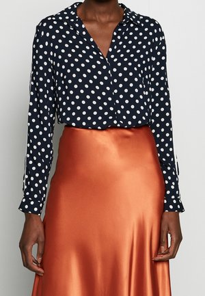 Persoon draagt een marineblauw shirt met lange mouwen met witte stippen en een glanzende, koperkleurige satijnen rok.