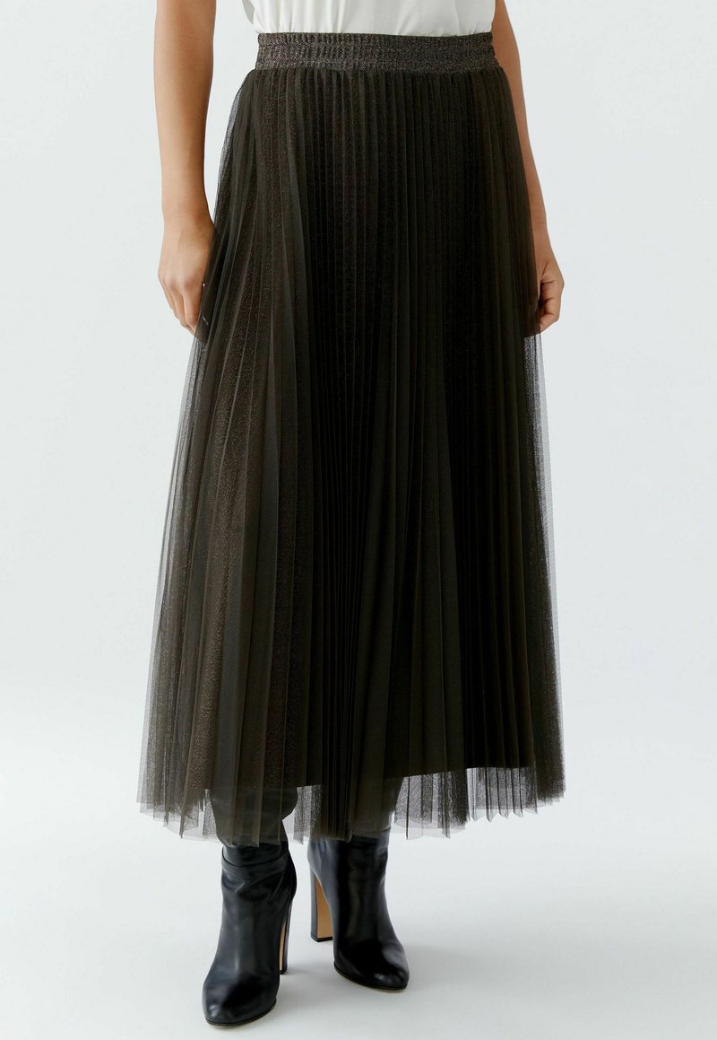 Pleated, ankle-length tulle skirt in dark green with a smocked waistband. Styled with black heeled boots for contrast.