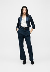 Femme debout, les mains dans les poches, portant un blazer court bleu marine, un chemisier en satin blanc, un pantalon large taille haute et des bottines noires.
