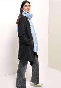 Cappotto nero con colletto, sciarpa blu testurizzata, jeans grigi a gamba larga e scarpe bianche con dettagli neri. Modella in piedi su sfondo chiaro.