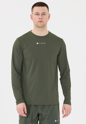 Homme portant une chemise à manches longues vert olive et un short avec le logo "VIRTUS", debout devant un fond clair uni.