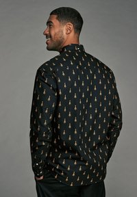 Chemise noire à boutons avec un motif d'arbres dorés en répétition. Le tissu semble lisse, avec des manches longues et un col standard.