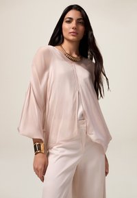 Lichtroze, luchtige blouse met vloeiende vleermuismouwen en een knoopsluiting aan de voorkant. Gekocht met crèmekleurige broeken en een gouden statement ketting.