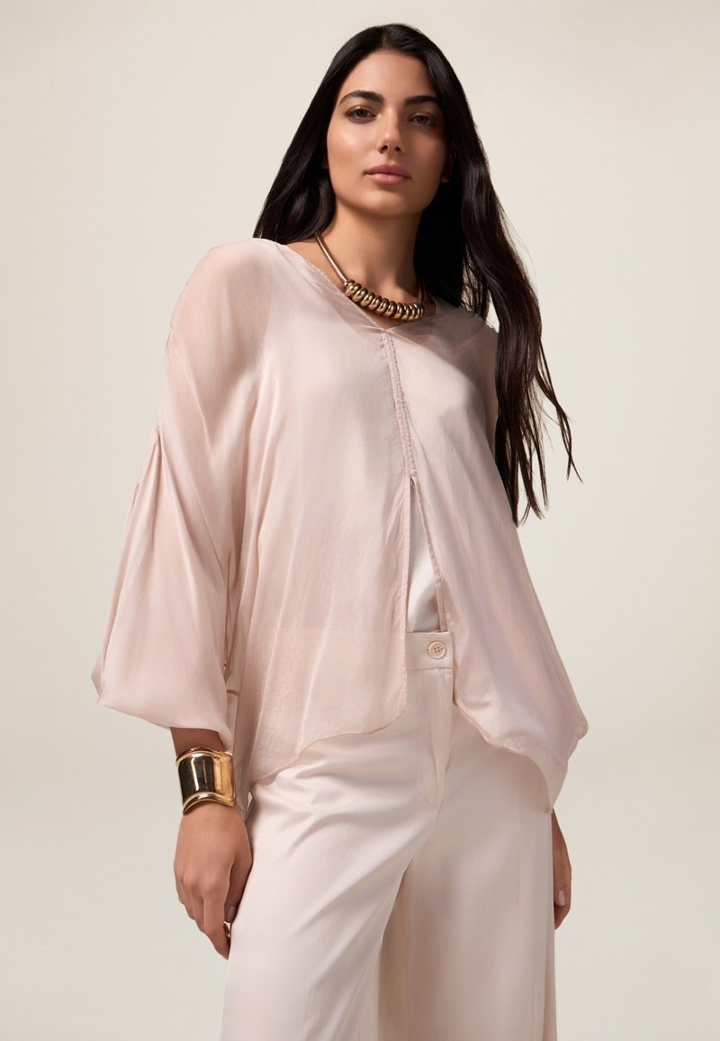 Lichtroze, luchtige blouse met vloeiende vleermuismouwen en een knoopsluiting aan de voorkant. Gekocht met crèmekleurige broeken en een gouden statement ketting.