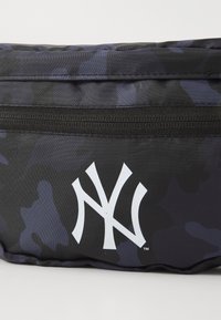 Sac en nylon avec un motif camouflage noir. Présente un logo "NY" blanc et une fermeture éclair noire le long de la couture supérieure.