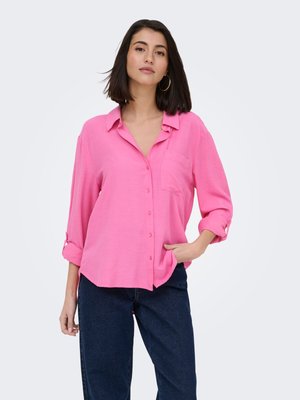 Mujer con cabello oscuro hasta los hombros, que lleva pendientes de aro dorados, una camisa rosa suelta abotonada y vaqueros azul oscuro, de pie con una mano en el bolsillo.