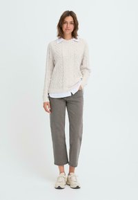 Suéter de punto trenzado color beige claro, camisa blanca con cuello por debajo, pantalones rectos grises y zapatillas beige con suelas texturizadas.