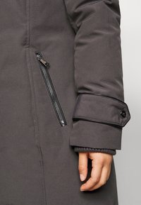 Närbild av en grå kappa med en knäppt remmudd och en vertikal dragkedjeficka nära handleden, hand delvis synlig.