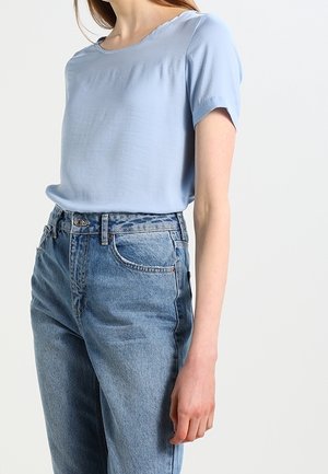 Femme portant un chemisier bleu clair à manches courtes, rentré dans un jean bleu taille haute, debout devant un fond uni.