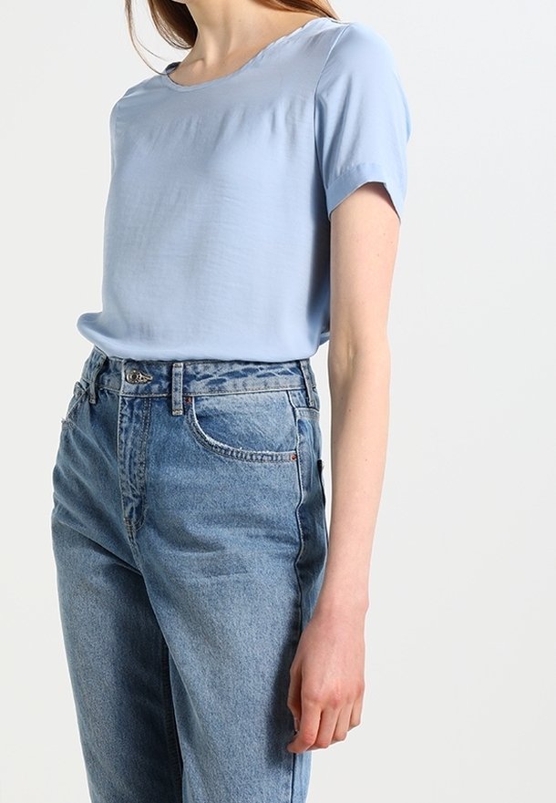 Femme portant un chemisier bleu clair à manches courtes, rentré dans un jean bleu taille haute, debout devant un fond uni.