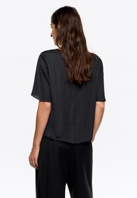 Czarna bluzka z krótkim rękawem o luźnym kroju i gładkiej fakturze, z prostym dołem i subtelnym połyskiem, noszona z dopasowanymi czarnymi spodniami.