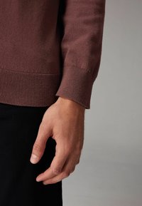 Barna kötött pulcsi mandzsetta bordázott textúrával, fekete anyag fölött viselve. Egy emberi kéz pihen a közelben, természetes bőrszínt és vonásokat mutatva.