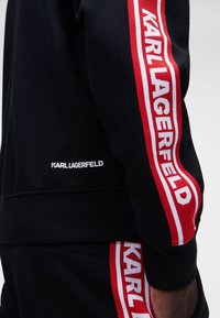 Sudadera negra con cinta del logo "KARL LAGERFELD" en rojo y blanco en las mangas, de textura suave y ajuste ceñido.