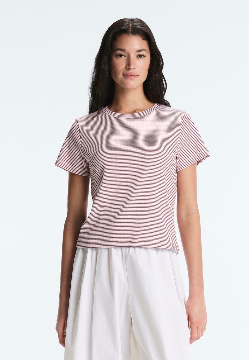 Femme aux longs cheveux foncés portant un t-shirt à manches courtes rayé rose et blanc et un pantalon blanc ample, debout devant un fond uni.