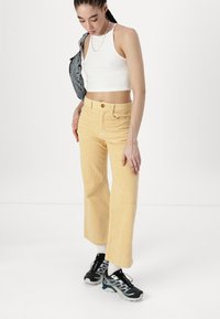 Pantaloni larghi in corduroy giallo con sottili righe verticali, chiusura con bottone e tasche frontali. Abbinati a un top corto bianco e sneakers.