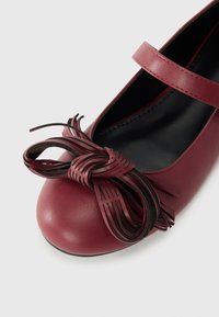 Burgundin nahkainen ballerinatossu pyöreällä varpaalla, jossa on solmittu nauha mustilla raidallisilla yksityiskohdilla ja ohut nilkkaremmi.