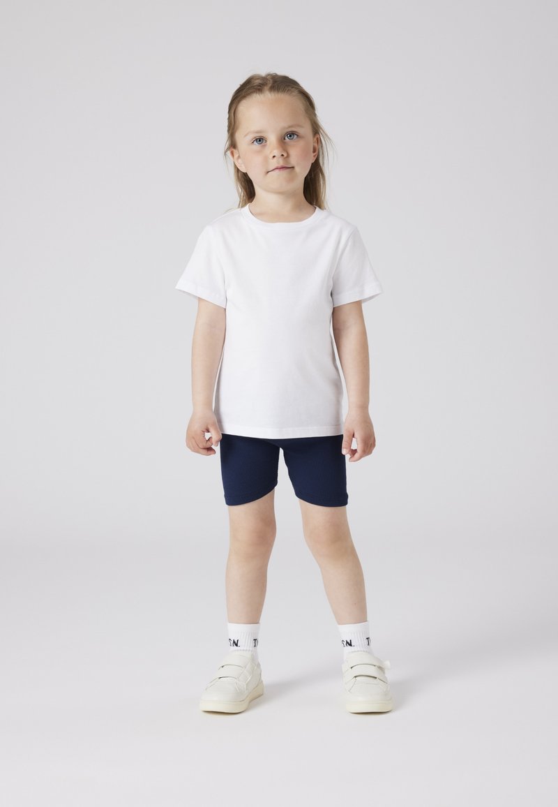 T-shirt en coton blanc à manches courtes associé à un short bleu marine. L'enfant porte des baskets blanches avec des scratchs et des chaussettes à motifs.