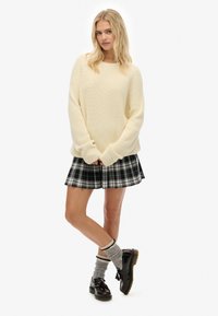 Giovane donna che indossa un maglione di maglia color crema, una gonna plissettata a quadri neri, calze grigie e mocassini marroni scuri, in piedi con le gambe incrociate.