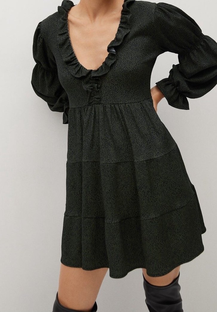 Robe noire à manches longues avec un col en V profond à volants et une jupe à étages, portée avec des bottes noires montant jusqu'aux genoux, présentée sur un fond uni.