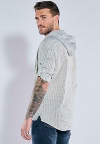 Leicht gestreiftes Hemd mit Knopfleiste und Kapuze in hellen Farben. Mit hochgekrempelten Ärmeln und abgerundetem Saum. Getragen mit dunkelblauen Jeans.