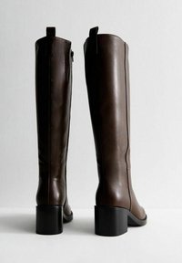 Stivali alti in pelle marrone con un design elegante, cerniera laterale e tacco a blocco. La texture è liscia e lucida, con una silhouette minimalista.