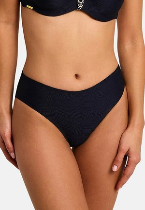 Femme portant un bas de bikini noir texturé et un haut assorti, debout, les mains détendues le long du corps sur un fond blanc.