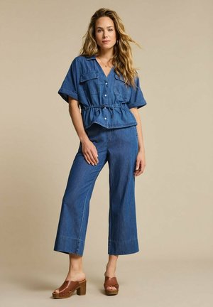 Vrouw staat met hand op heup, draagt een blauw denim kortärmelig overhemd met trekkoord en wijde jeans met bruine sandaalhakken.