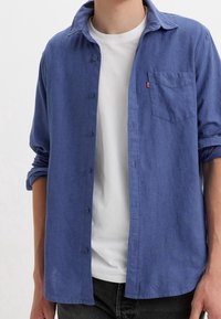 Niebieska koszula na guziki wykonana z lekkiego materiału, z kołnierzem, pojedynczą kieszenią na piersi i podwiniętymi rękawami. Noszona na białej koszulce.