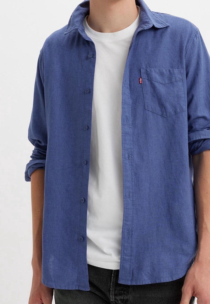 Niebieska koszula na guziki wykonana z lekkiego materiału, z kołnierzem, pojedynczą kieszenią na piersi i podwiniętymi rękawami. Noszona na białej koszulce.