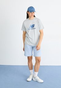 Light gray t-shirt with blue logo, light blue shorts, and denim cap. Footwear is white with blue accents. Minimalist design and casual style.