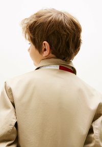 Veste beige avec un col, présentant un accent rouge sur la doublure intérieure. Texture douce et finition lisse, adaptée à une tenue décontractée.