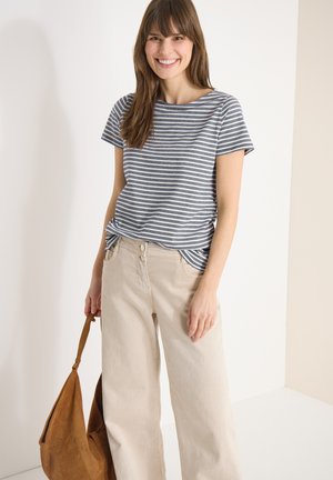 Femme souriante portant un t-shirt à manches courtes rayé et un pantalon beige à jambes larges, tenant un sac à bandoulière en cuir marron devant un fond uni.