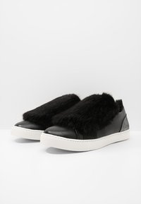 Pár černých slip-on tenisek s bílými podešvemi a huňatým černým svrškem zakrývajícím tkaničky, na jednoduchém bílém pozadí.