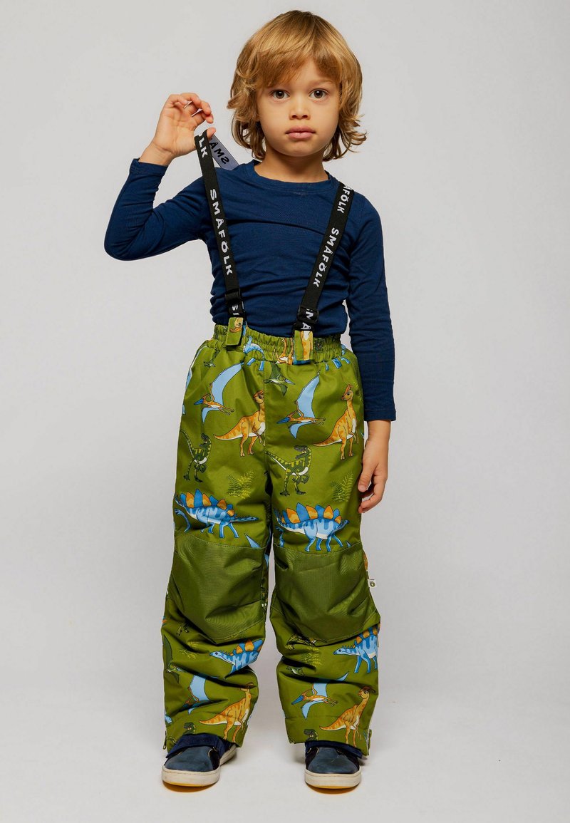 Enfant aux cheveux blonds portant une chemise bleu marine à manches longues et un pantalon de neige vert avec un imprimé dinosaure et des bretelles noires, debout devant un fond uni.