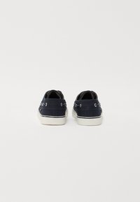 Zapatos de lona azul marino con suelas de goma blanca. Las características incluyen ojales de metal y una parte superior texturizada. La vista trasera revela costuras y un acento de logo.