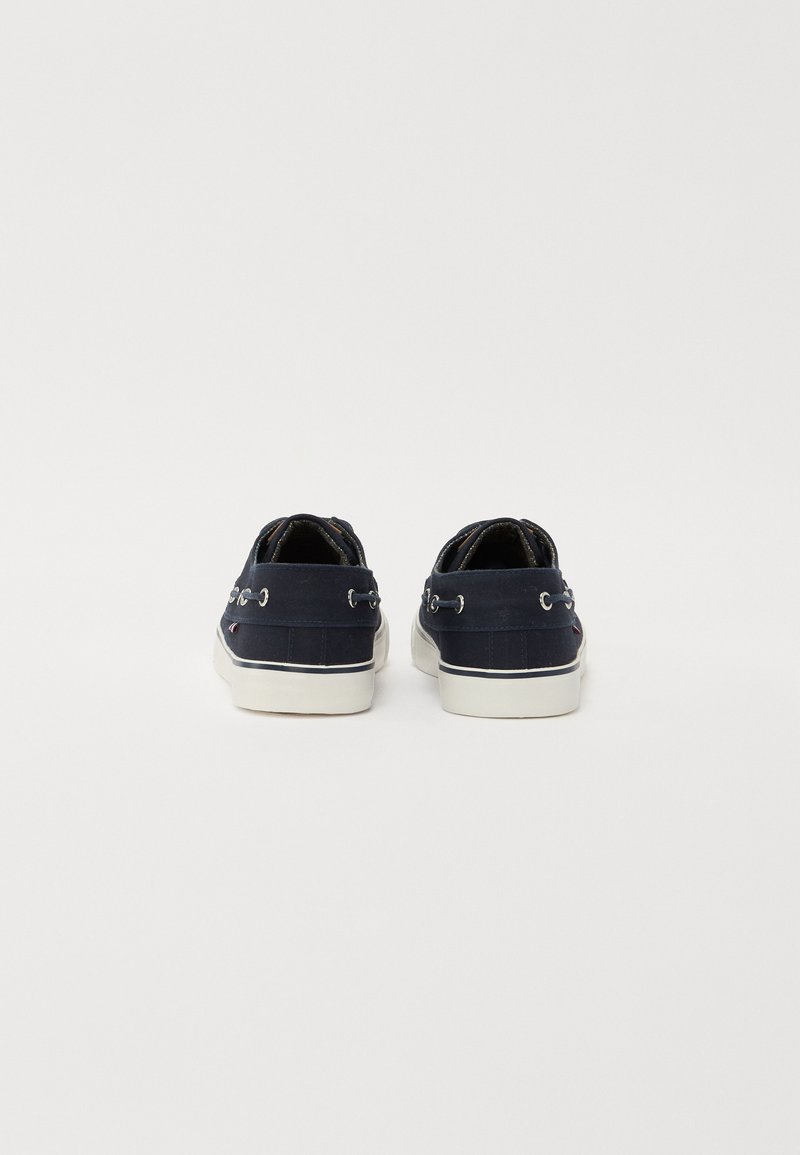 Zapatos de lona azul marino con suelas de goma blanca. Las características incluyen ojales de metal y una parte superior texturizada. La vista trasera revela costuras y un acento de logo.
