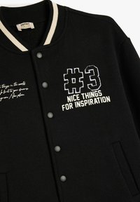 Svart sweatshirtjacka med beige randig krage, tryckknappar, broderad text "#3 FINA SAKER FÖR INSPIRATION" och ytterligare vit textdetalj.
