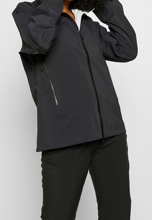 Personne portant une veste noire zippée avec des poches latérales zippées et un pantalon noir, ajustant la capuche ou le col, sur un fond uni.