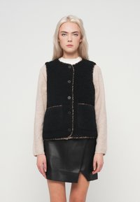 Μαύρο γιλέκο από φλις με υφή, κλείσιμο με κουμπιά, δύο μπροστινές τσέπες και αντίθετη διακόσμηση, φορεμένο πάνω από ένα ανοιχτόχρωμο τοπ και δερμάτινη φούστα.