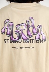 Beige t-skjorte med et glansfullt, flerfarget «ALPHA» grafikk i lilla og svart over teksten «STUDIO EDITION» i fet svart skrift.