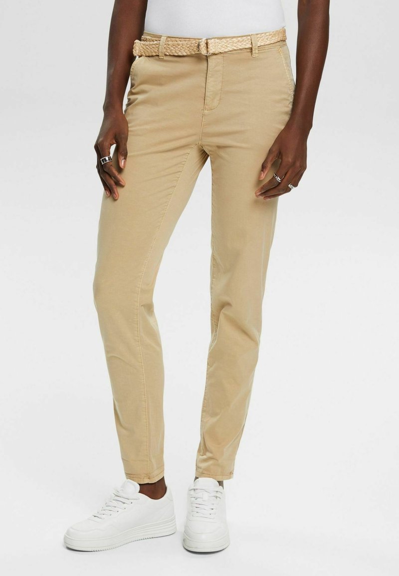 Personne portant un pantalon beige slim avec une ceinture tressée et des baskets blanches, debout avec les mains détendues le long du corps.