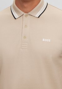 Polo beige en tissu texturé, doté d'un col à rayures blanches et noires, d'une patte de boutonnage à trois boutons et d'un logo "BOSS" brodé.