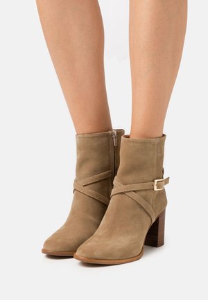 Kaki mockaskinn ankelskor med spetsig tå, korsrembetoning, sidodragkedja och en staplad träklack. Slät textur, minimal detaljer.