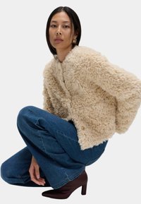 Beige Kunstpelzjacke mit weicher, strukturierter Oberfläche, die eine lässige Form und zwei goldene Knöpfe aufweist. Kombiniert mit blauen Jeans mit weitem Bein und braunen High Heels.