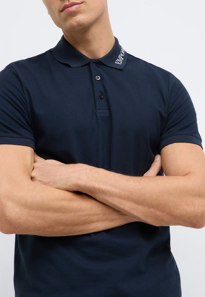 Tamsiai mėlynas polo marškinėlis su tekstūruota medžiaga, trijų mygtukų užsegimu ir apykakle su siuvinėtu prekės ženklo pavadinimu ant apykaklės.