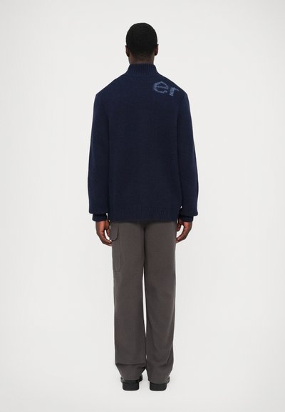 Marinblå stickad tröja med hög krage, prydd med en diskret logodetalj på baksidan, ihop med grå cargobyxor och svarta skor.