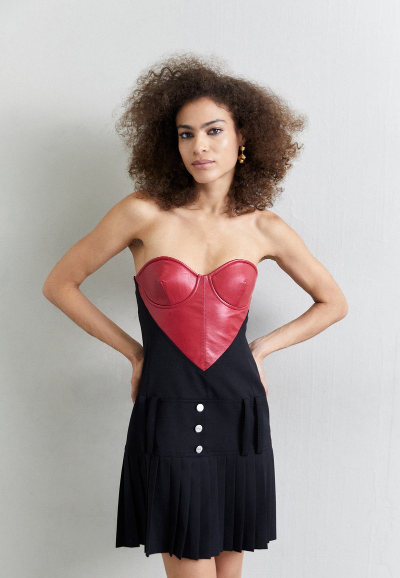 Μαύρο φόρεμα με εφαρμοστό, κόκκινο δερμάτινο κορσάζ σε σχήμα καρδιάς, που διαθέτει πλαϊνές τσέπες και πλισέ φούστα με τρία λευκά κουμπιά στη μέση.