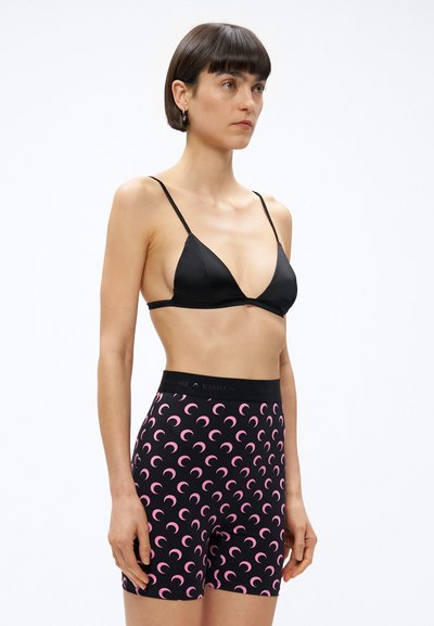 Kobieta z krótkimi ciemnymi włosami, ubrana w czarny trójkątny biustonosz i czarne szorty z wysokim stanem zdobione różowymi wzorami w kształcie półksiężyca, stoi na białym tle.