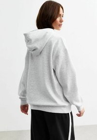 Gray hoodie with a spacious hood, long sleeves, and ribbed cuffs. Soft, textured fabric and a relaxed fit. Worn with black pants featuring white stripes.