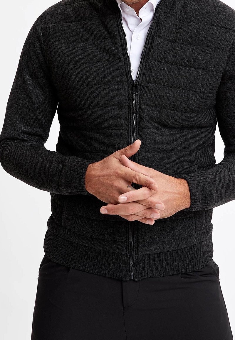 Czarna kurtka na zamek z pikowanymi panelami, z prążkowanym kołnierzem i mankietami, wykonana z teksturowanej tkaniny, dopasowany fason i ukryte kieszenie.